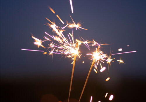 Eine Hand halt zwei brennende Wunderkerzen vor einem abendlich dunkelblauen Himmel.